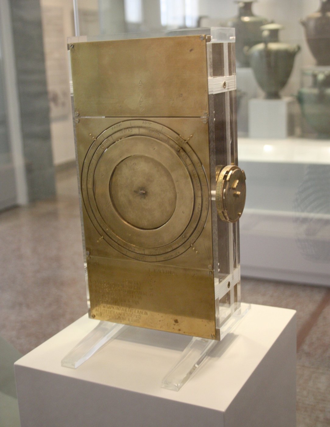 Figure 7 — A modern reconstruction of the mechanism on display in the National Archaeological Museum, Athens, near the original fragments: a bronze-finished case with the front dial at the centre, inscribed p…