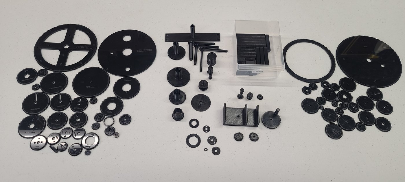 Figure 4 — The parts of an Open Antikythera kit laid out: black laser-cut acetal gears, including the large four-spoked main wheel, 3D-printed brackets and the crank, and a small clear acrylic sub-assembly.