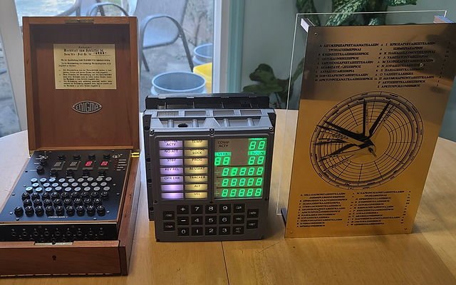 Figure 2 — S&T GeoTronics' three replicas side by side: the Open Enigma in its wooden case, the Open DSKY with its lit keypad and displays, and the Open Antikythera's gold-finished front plate with its needles.