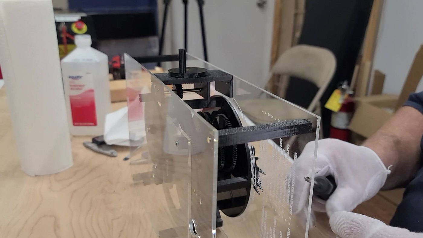 Figure 3 — An UpFront unit in final assembly at S&T's workshop: the clear acrylic plates with their engraved lettering, black acetal gears behind them, the 3D-printed crank block on top, and the assembler in …