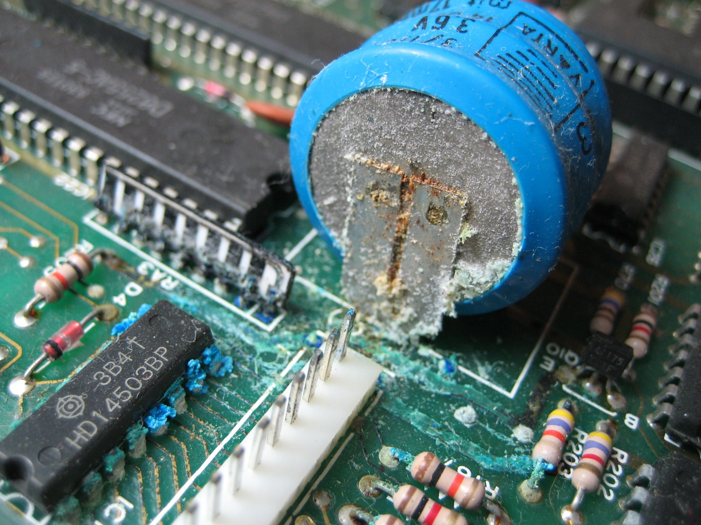 Figure 5 — Corrosion spreading from a leaking component across traces and chip leads, on a 1980s synthesizer board. Apple-1s have no batteries, but the same green-white deposits appear wherever moisture, salt…