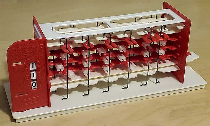 Figure 1 — An original E.S.R. Digi-Comp I with a program in place: logic tubes on the front pegs of the three red flip-flops, the six logic rods hanging across them, and the read-out at the left showing one b…