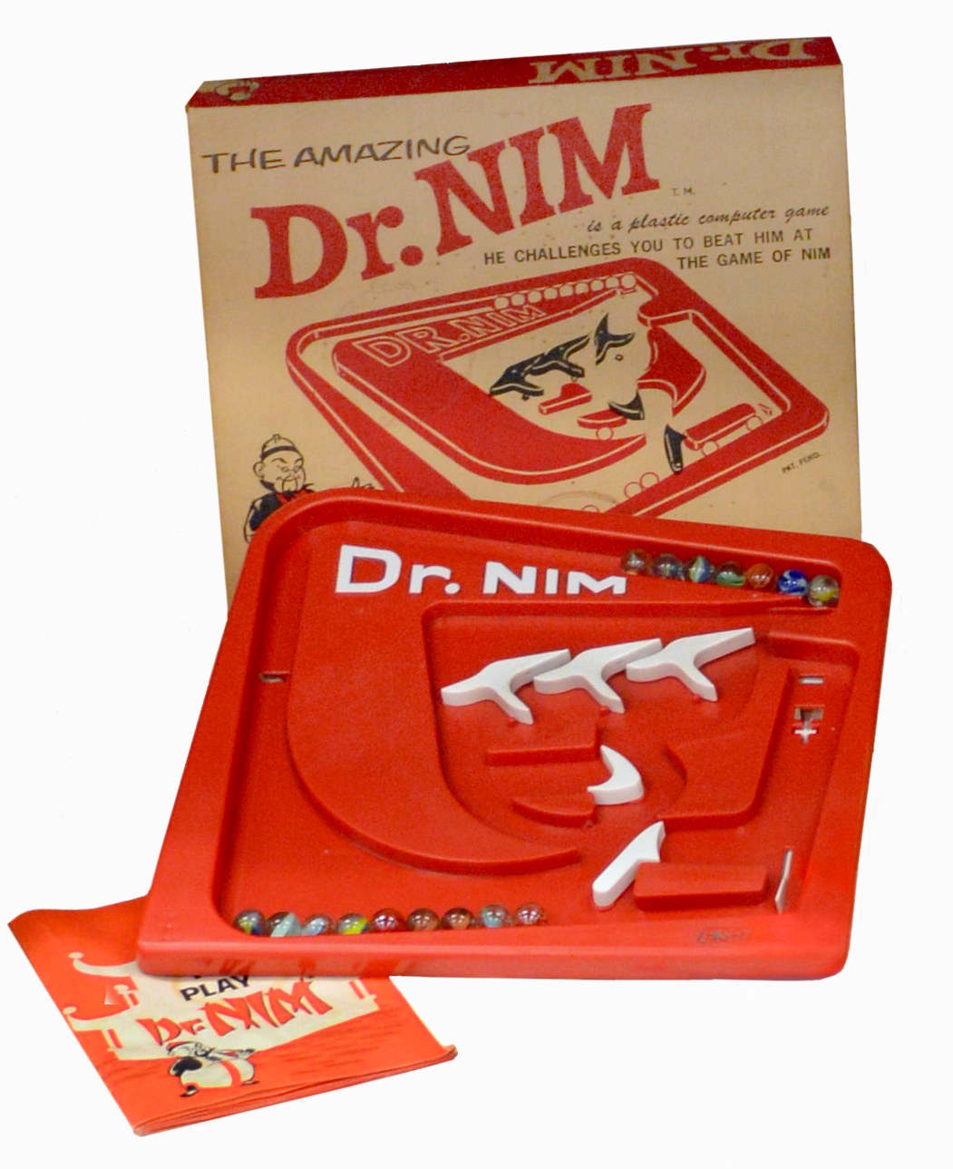 Figure 4 — The Amazing Dr. Nim, with its box and the "Play Dr. Nim" instruction booklet. The marbles wait in the rack across the top; the three white flip-flops in the middle are the machine's memory; the tur…