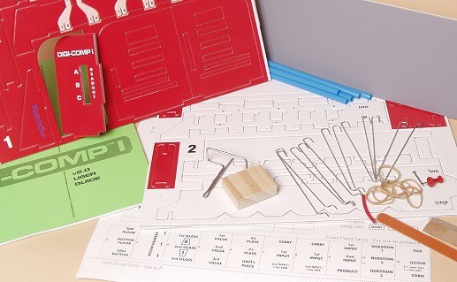 Figure 3 — The Digi-Comp I v2.0 kit laid out before assembly: printed and laminated board sheets with the plates and frame parts, the pre-bent piano-wire rods, rubber bands, straws, a wooden crank base and th…