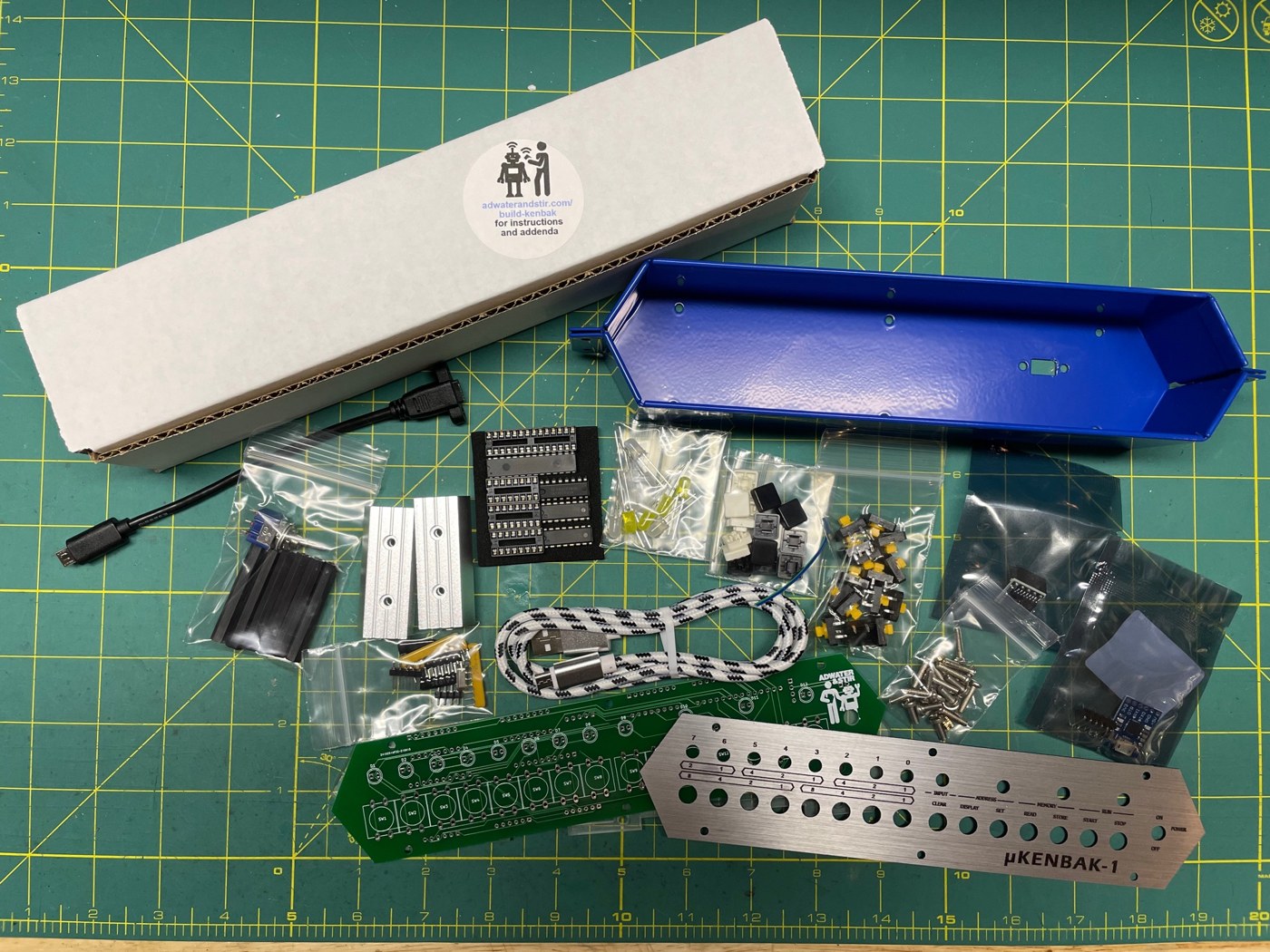 Figure 7 — The µKENBAK-1 kit as it arrives: the blue steel case and aluminium end pieces, the brushed-aluminium front panel, the circuit board, keyswitches and keycaps, LEDs and bagged components.