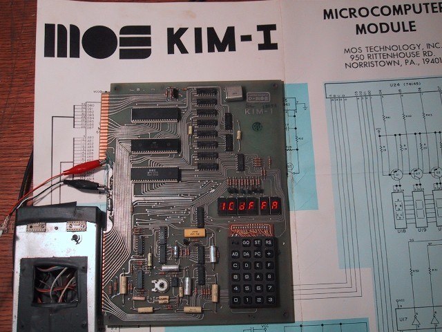 Figure 4 — A KIM-1 in use: power leads clipped to the lower edge connector, the display lit, and the board's schematic sheet behind it. The six digits show a four-digit address on the left and the two-digit c…