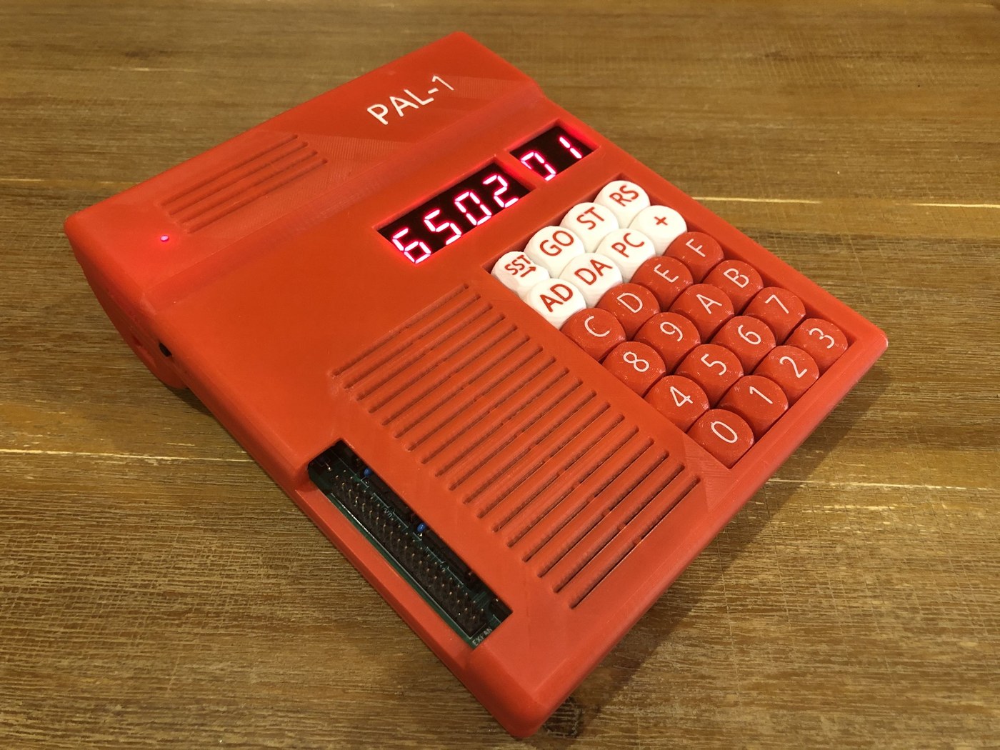 Figure 5 — A PAL-1 in a red printed case, powered up and showing "6502 01". The PAL-1, a compact single-RIOT clone descended from the Micro-KIM, was the most widely sold KIM-1 replica of the early 2020s; it s…
