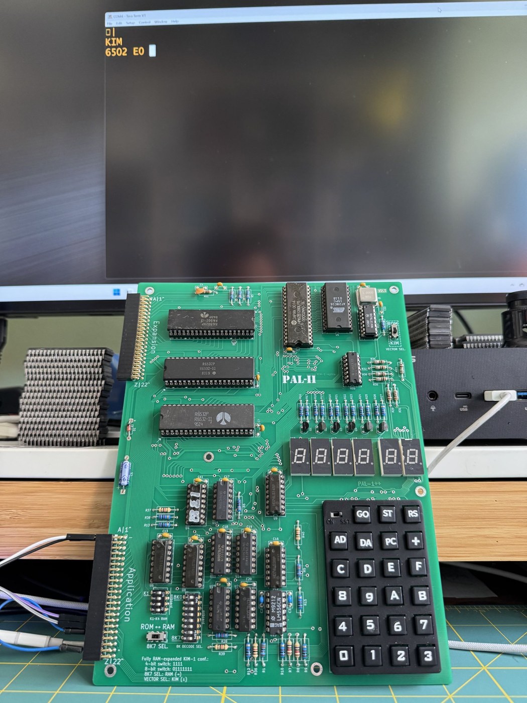Figure 1 — A finished PAL-2 at work: a USB-to-TTL adapter on the Application connector at lower left supplies both power and the serial link, and the terminal window behind shows the KIM monitor's prompt ("KI…