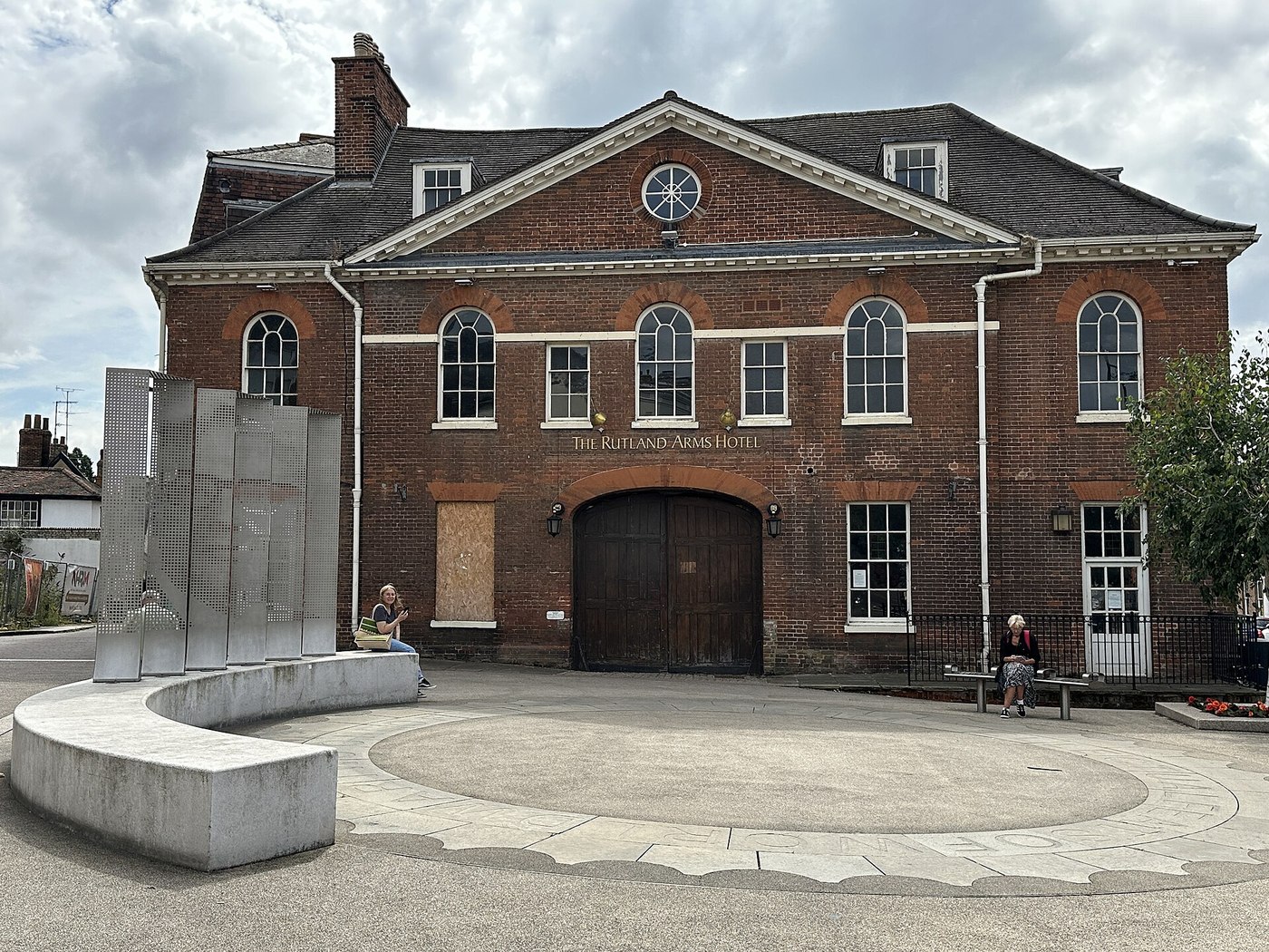 Figure 6 — The Bill Tutte memorial at Rutland Hill, Newmarket, photographed in 2024: steel panels and a circular paved base in front of the Rutland Arms, the hotel where Tutte's father once worked. The memori…