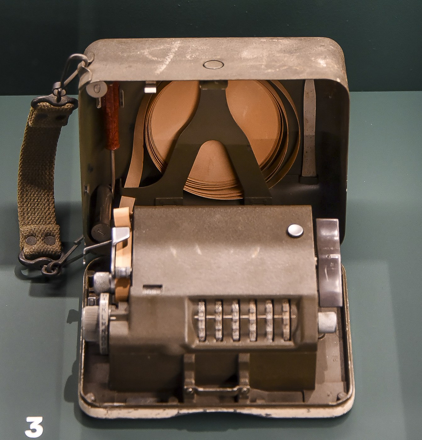 Figure 2 — An M-209 opened for use, with its webbing strap, in the Liberation Garden museum at Leopoldsburg, Belgium. The paper-tape spool sits in the lid; the drive knob is on the right and the setting knob …