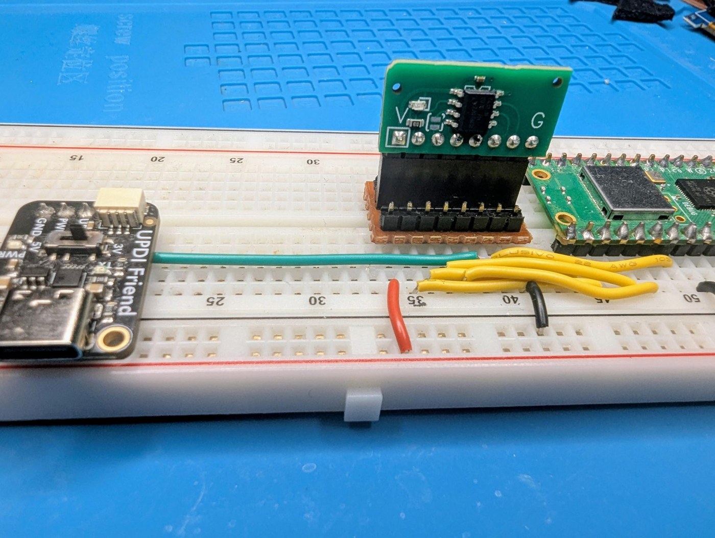 Figure 10 — The test jig: a card in the stripboard socket, plugged into a breadboard beside the Pico that runs the electrical checks and the Adafruit UPDI Friend that flashes it.