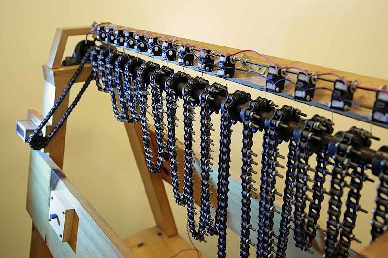Figure 6 — The reproduction's chain bank seen from one end: fifteen loops of roller chain hang from sprockets on the common shaft, long-tailed screws stand out from selected links, and a row of microswitches …